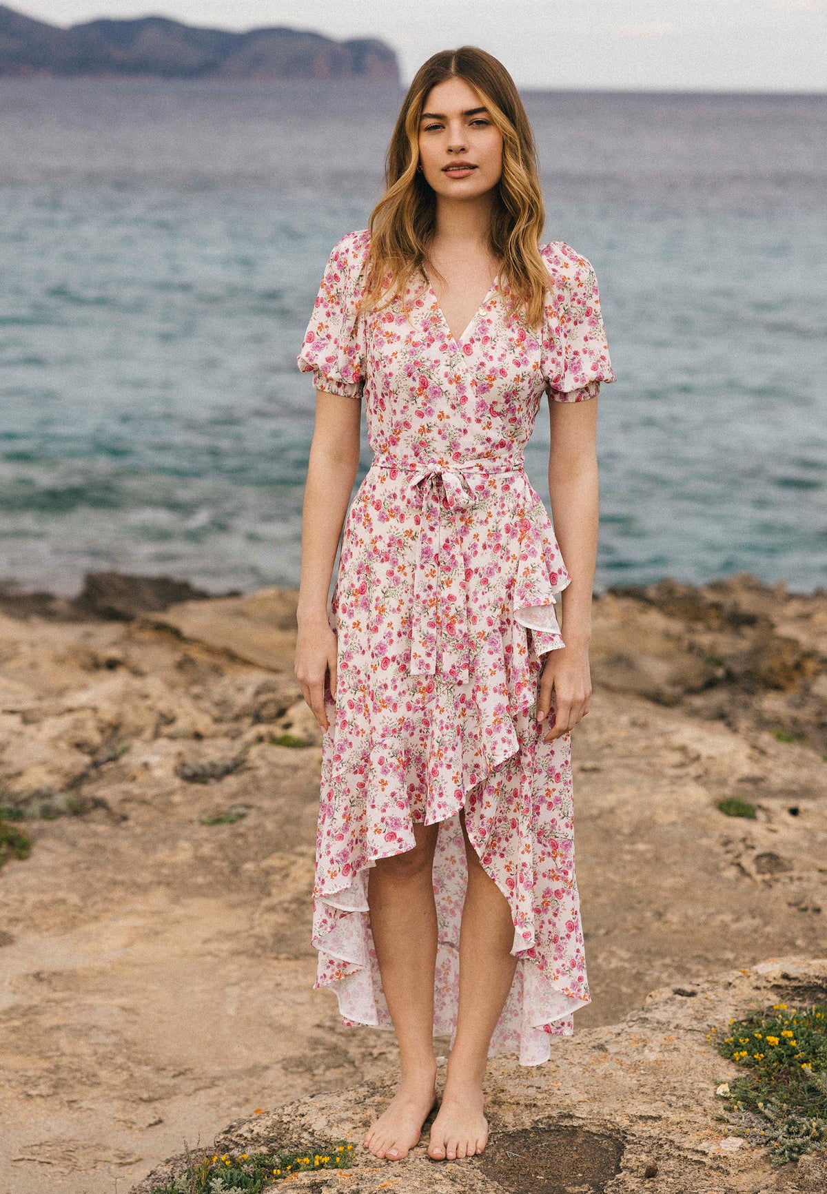 A woman in a pink and cream floral dress ISLAY stands barefoot on a rocky shore, with the calm sea in the background.
