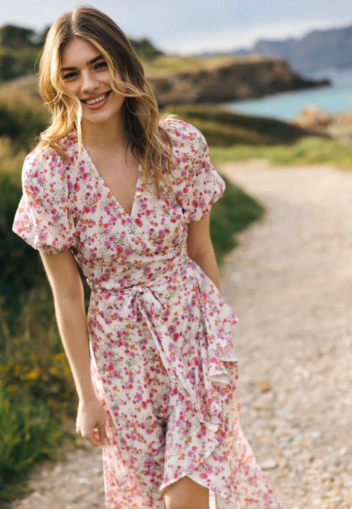A smiling woman in a floral short-sleeved envelope dress ISLAY walks along a coastal path, with water and rocks in the background.
