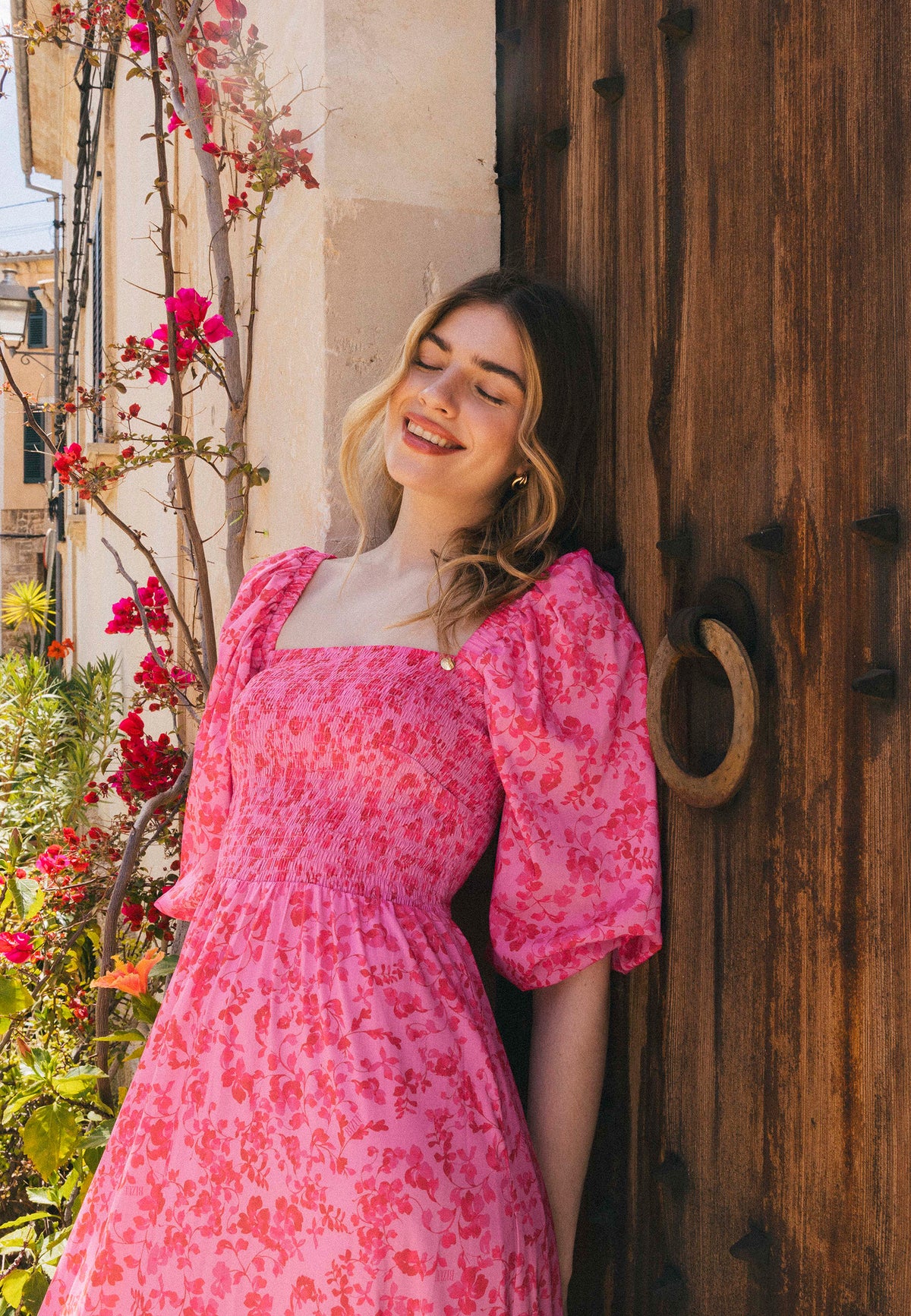 A woman in a pink floral dress LUMIA leaning against a wooden door, with her eyes closed and a smile on her face, surrounded by flowering vines.