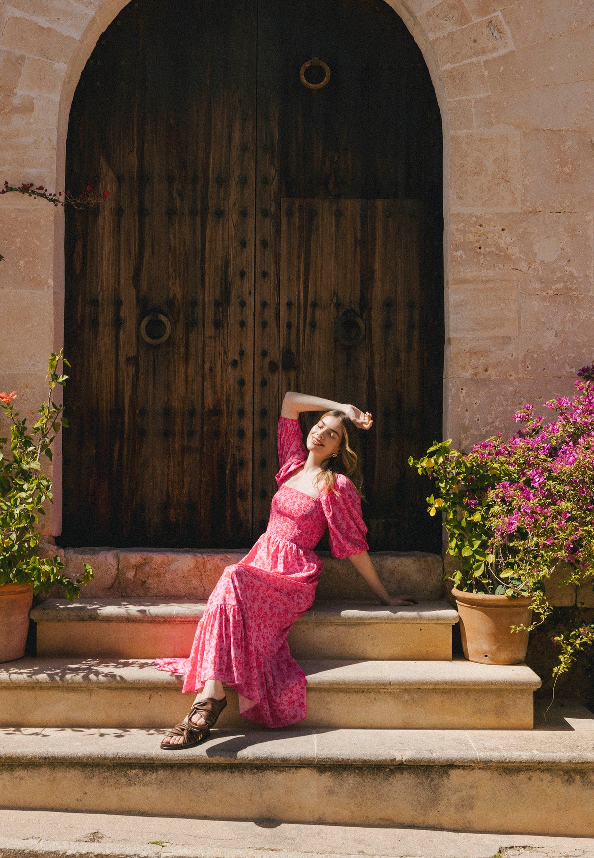 A woman in a pink dress sits on a stone staircase by a wooden door, surrounded by flower pots.