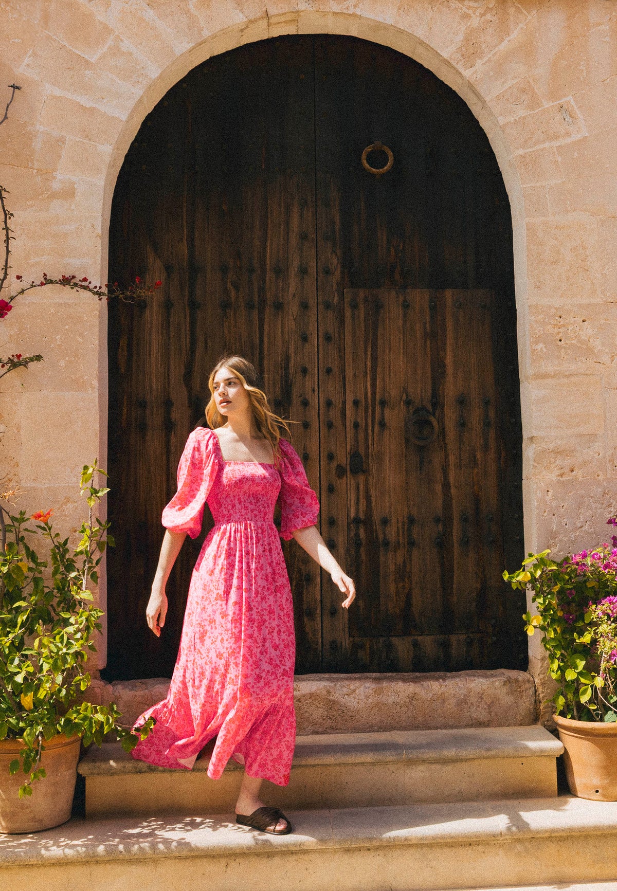 A woman in a pink floral LUMIA dress with buff sleeves stands in front of a large wooden door in a stone wall, surrounded by potted flowers.
