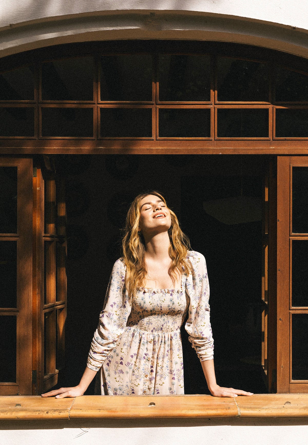 A model wearing a long PATRYA dress with a delicate floral pattern leans out of a wooden window, her face facing the sun with her eyes closed, in the light of the morning glow.