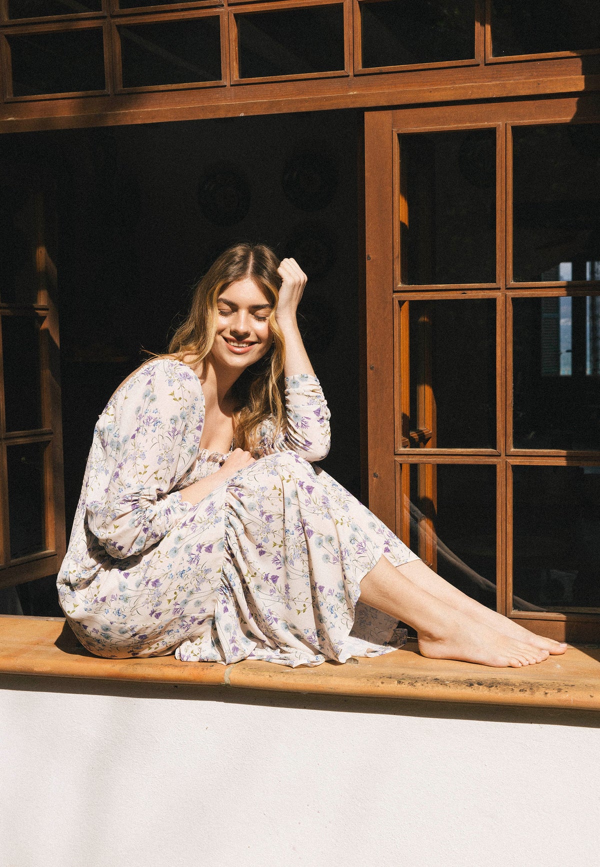 A model in a pastel PATRYA dress amidst the sunshine sits sideways on a wide window sill, with her head resting on her hand and her eyes closed.
