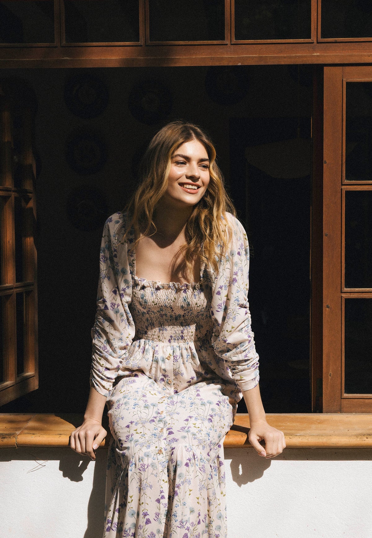 A smiling woman in a romantic PATRYA dress sits on the sill of an open window, enveloped in warm daylight, with her hair falling loosely on her shoulders.