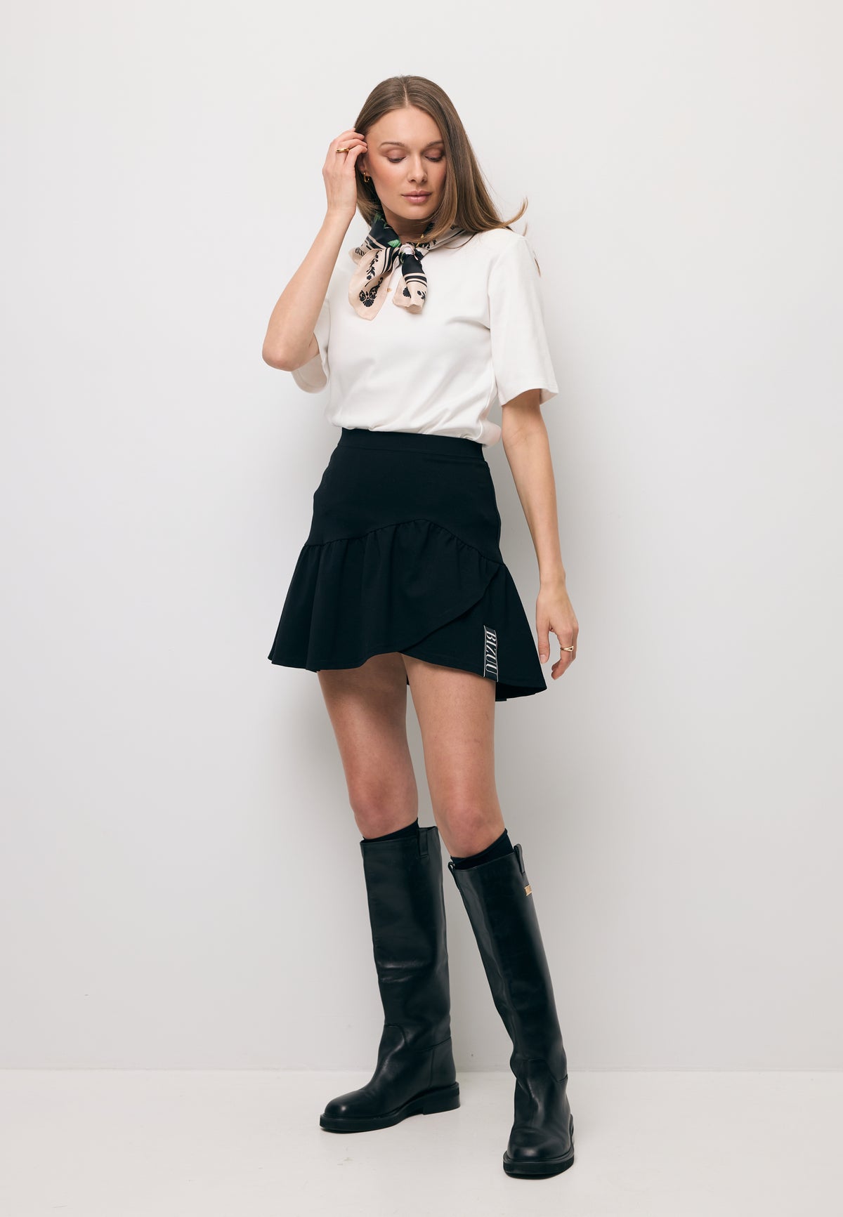The model against a white wall, wearing a cream MELISA T-shirt with a decorative logo and shoulder pads, paired with a black TULIAN mini skirt.