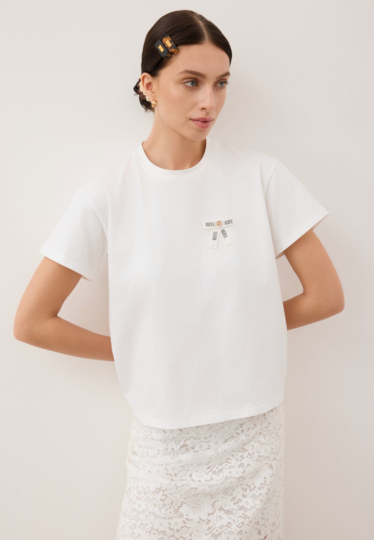 A model leaning against a white wall with her hands behind her back. She is wearing a white T-shirt with a TISHALLE logo bow and a white MILKY lace skirt.