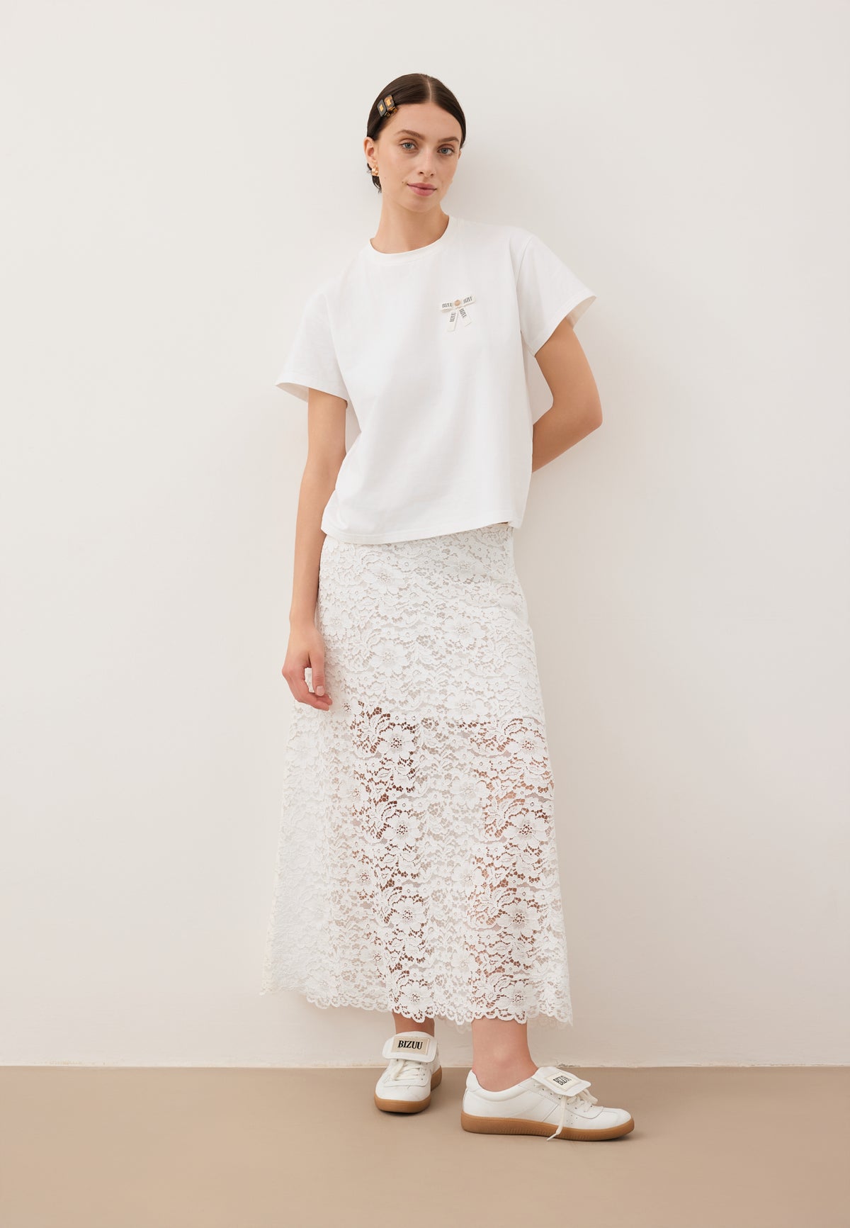 A model sitting on a sofa with her legs on the seat. She is wearing a white MILKY skirt, a T-shirt with a TISHALLE logo bow, and MOBBY shoes.