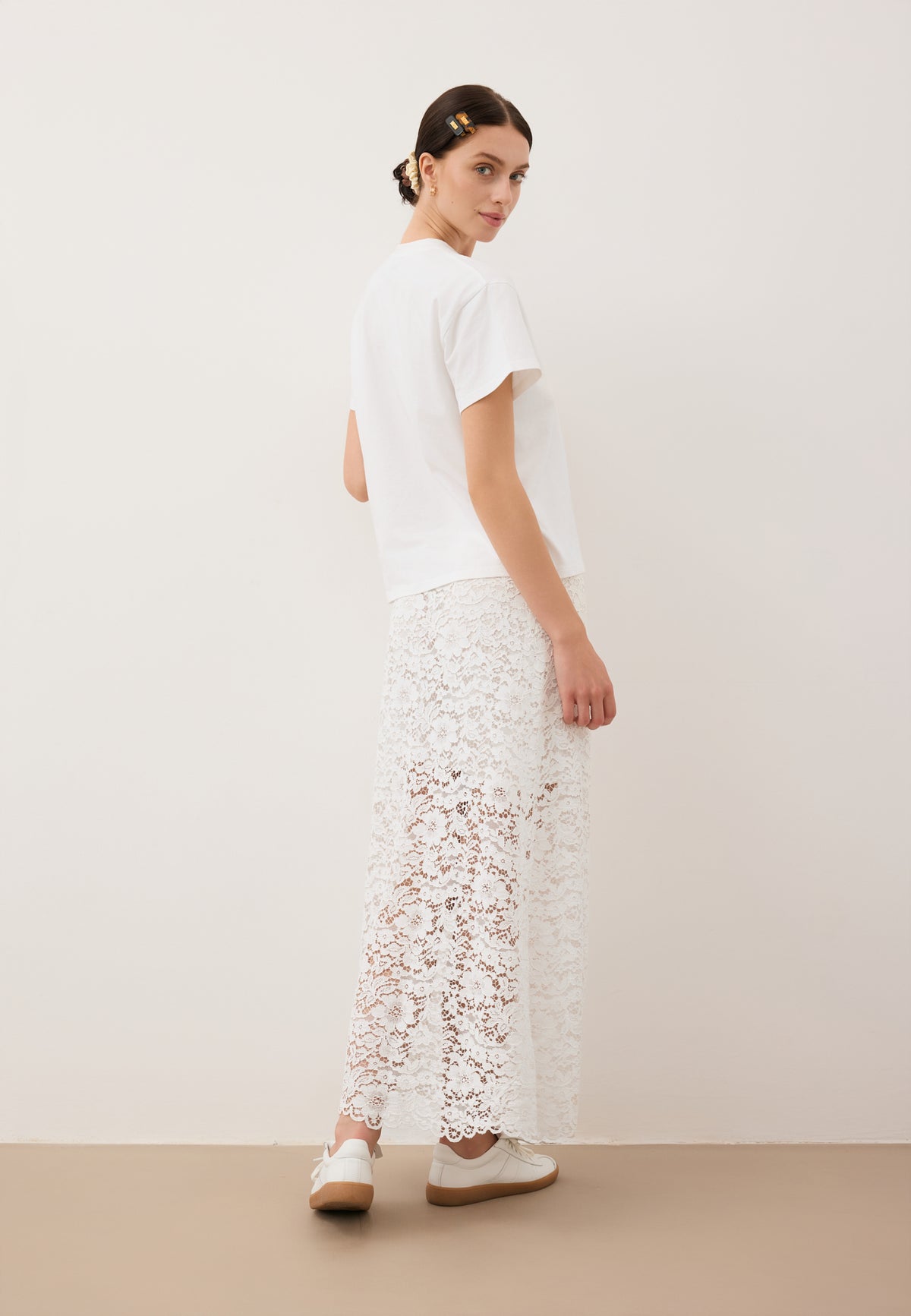 Model standing with her back to the camera, wearing a white MILKY lace skirt and a white TISHALLE T-shirt.