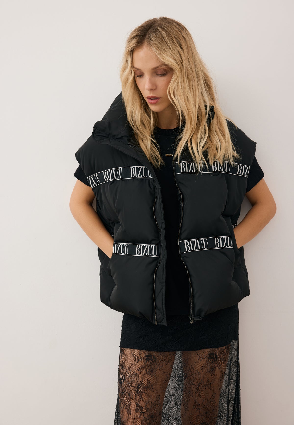 Model with hands in the pockets of the black AYASE down vest with logo tape, styled with the FLACE lace mini skirt.