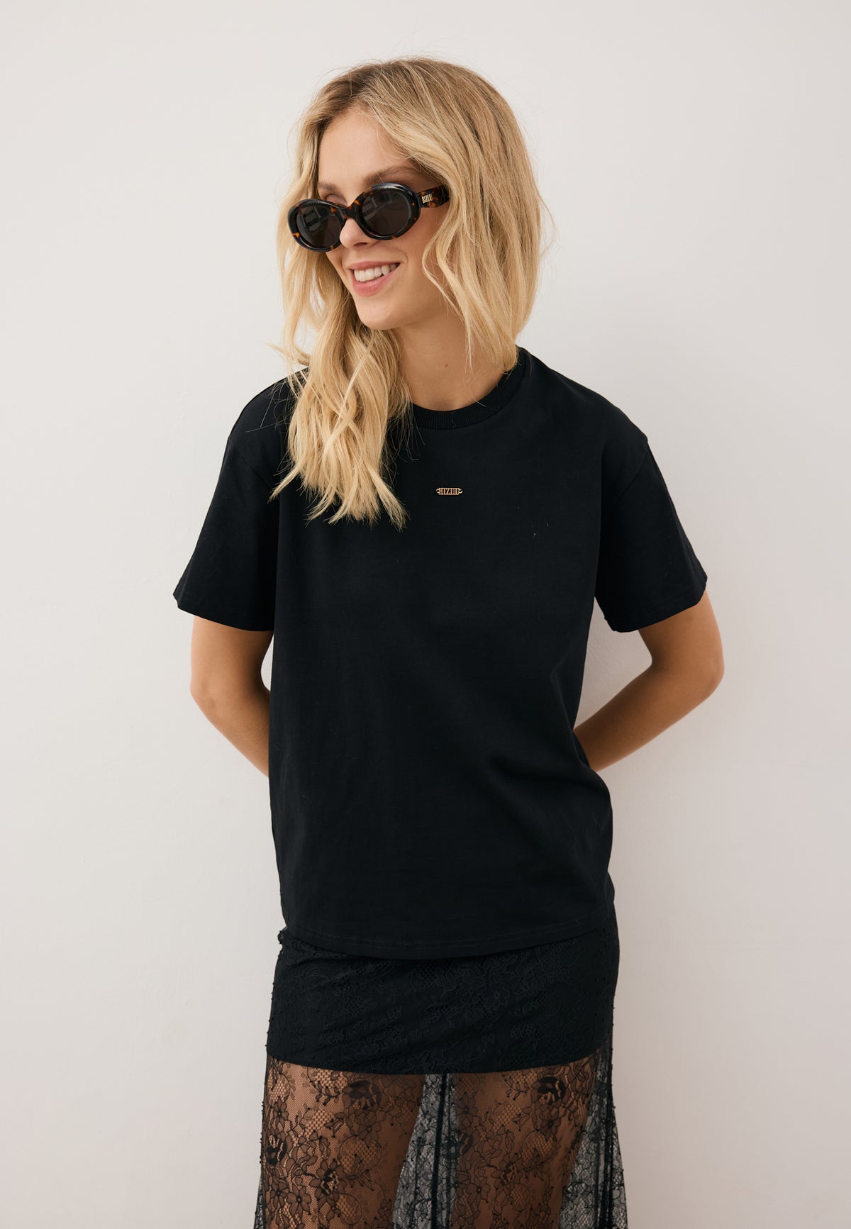 Model leaning against a wall, styled in the black TISHAL T-shirt, black lace FLACE skirt, and sunglasses.