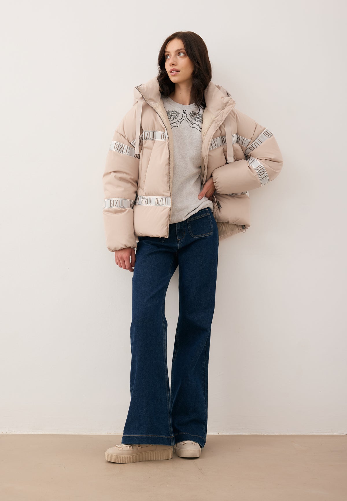 Model looking sideways with one hand on her hip, wearing an unzipped TOKYO down jacket, COLLIER sweatshirt, and DELPHI trousers.