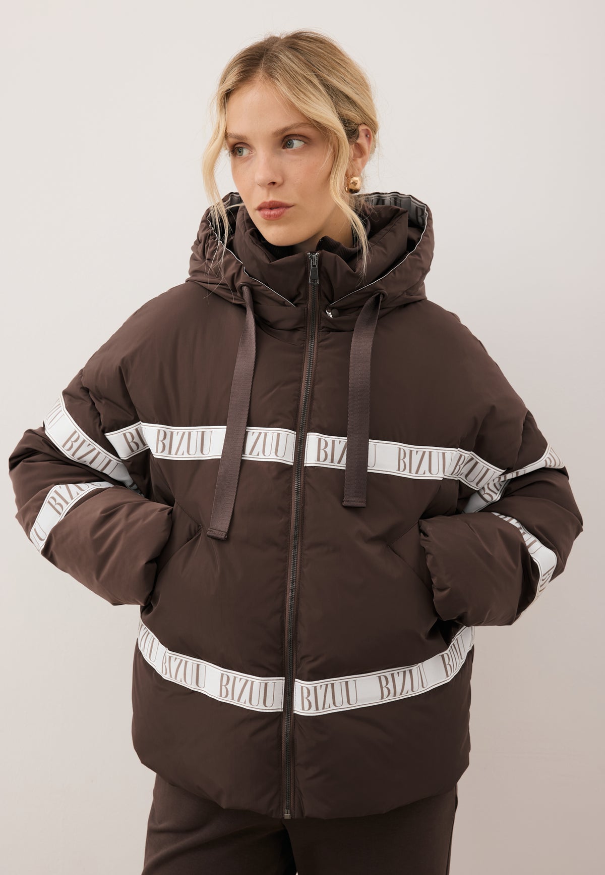 Close-up of the model with her hands in the pockets of the brown TOKYO jacket with natural filling and logo.