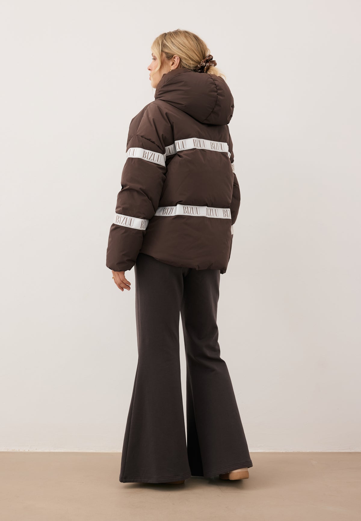 Model standing with her back turned in the brown TOKYO down jacket with branded tape and hood.