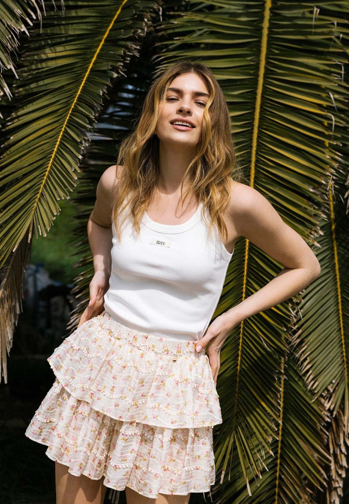 The model in a white EJRIS strapless top with BIZUU logo and COKO skirt with ruffles in a fine floral print poses against a background of palm leaves.