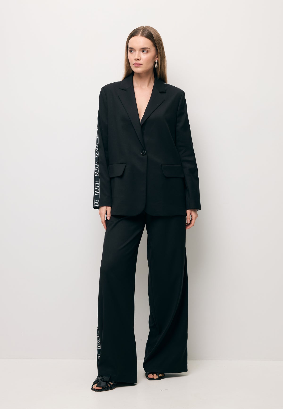 The model against a white wall, wearing a black suit with a white side stripe – the VANDA blazer and MICHAEL trousers.