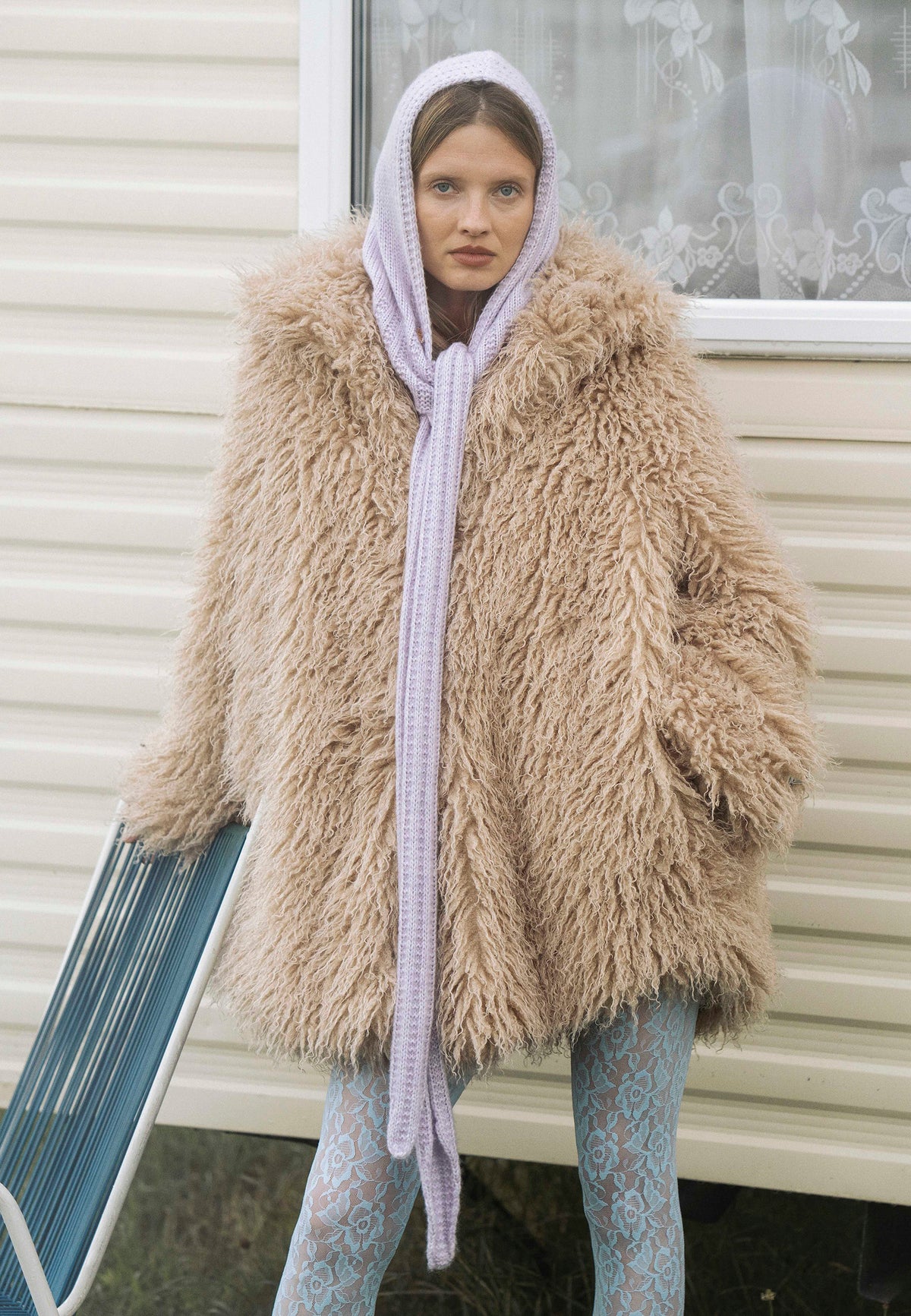 Model leaning against a chair in front of a house, dressed in the beige crimped long-hair VICENZA faux fur coat.