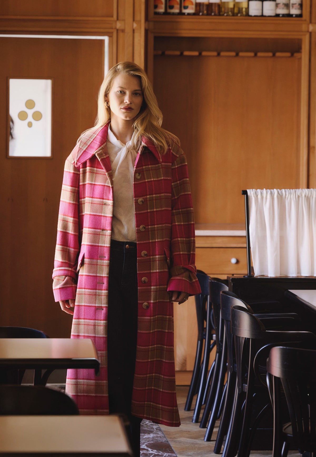 A model leaning against a white wall, wearing a pink WIVINA check coat and black NELLA pants.