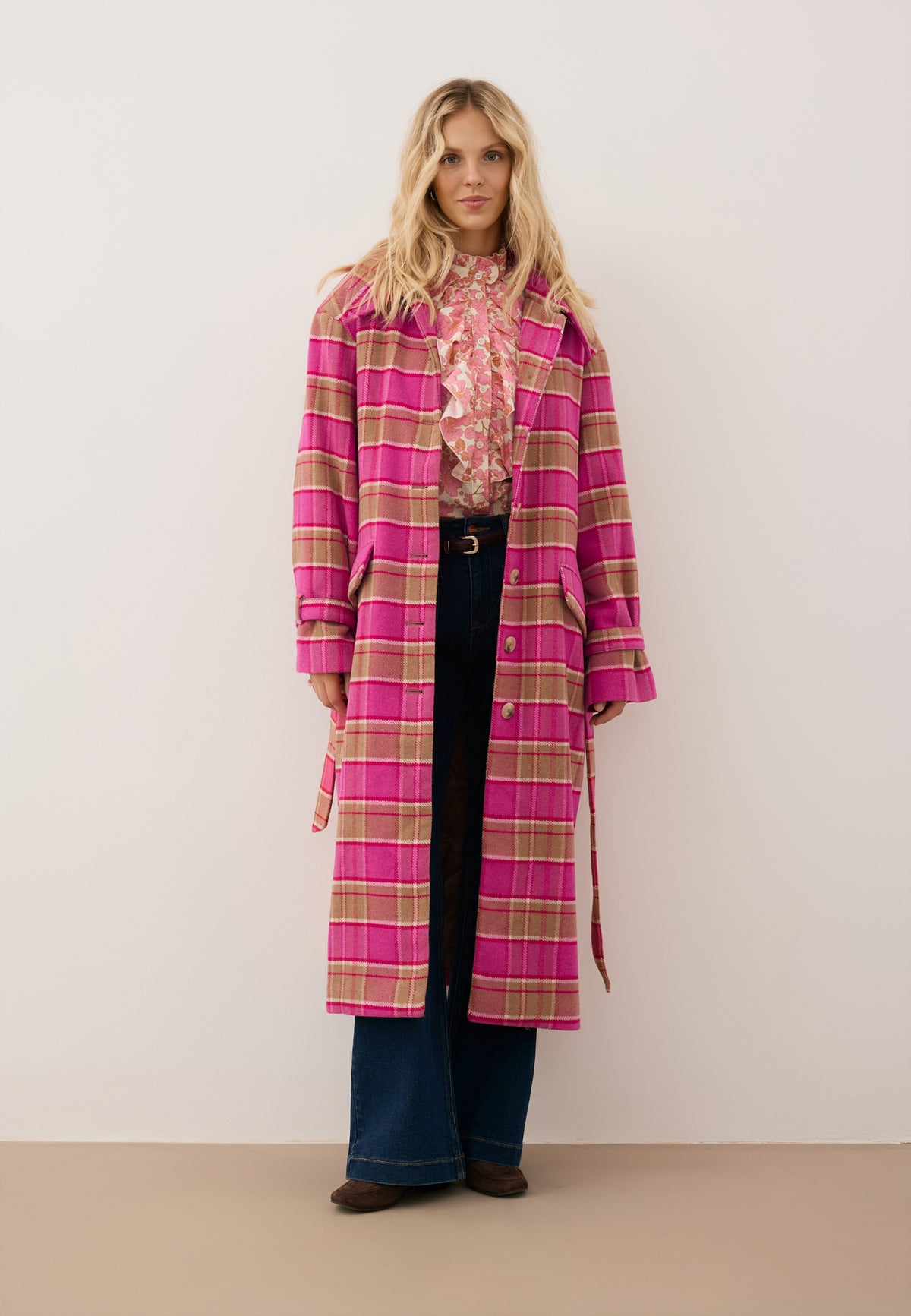 A model walking in a café against a wooden wall, wearing a pink WIVINA checkered coat, a DEFIORI bodysuit, and navy blue DELPHI pants.