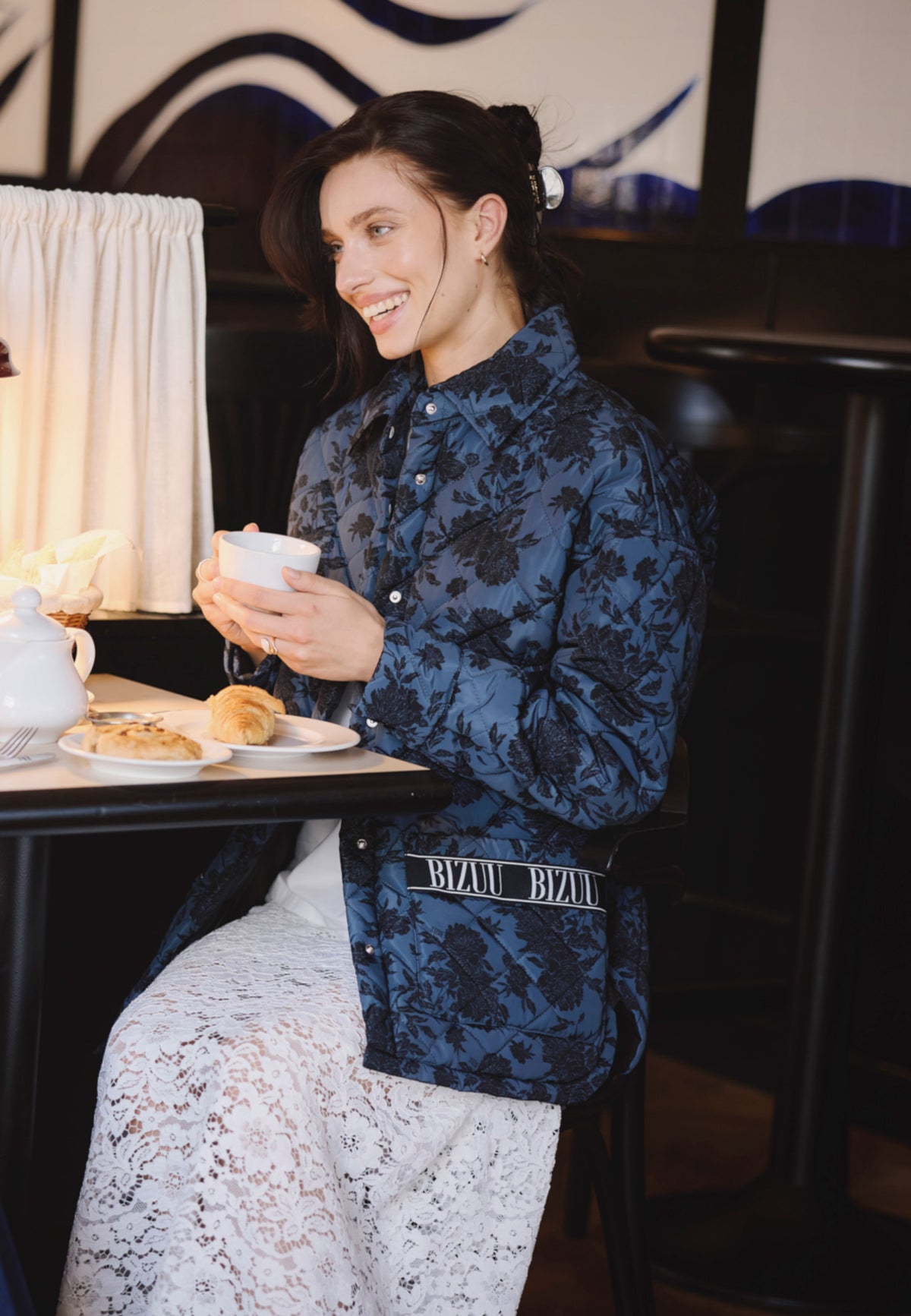 A model sitting at a table in a café. She is wearing a MARIN quilted jacket with a print and a white MILKY lace skirt.