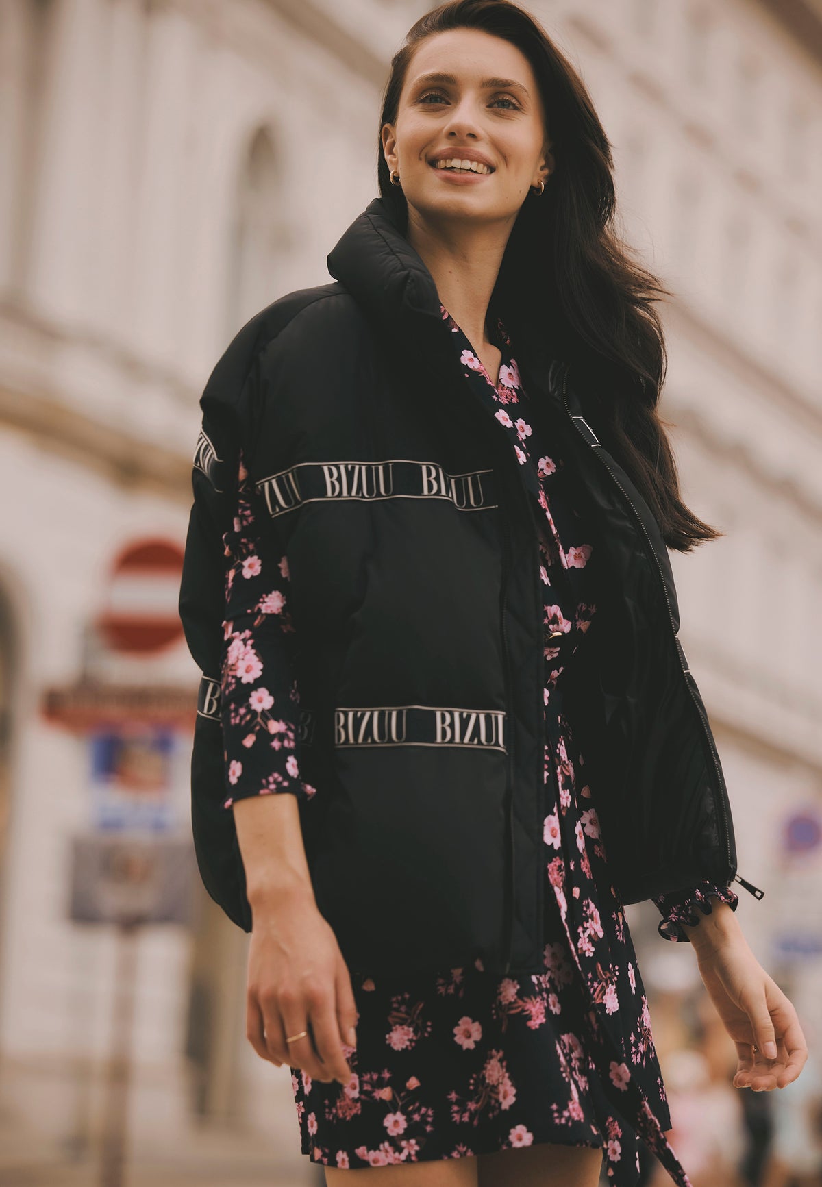 Smiling model standing on the street, wearing the black AYASE down vest with logo tape and the floral GEORGINA dress.