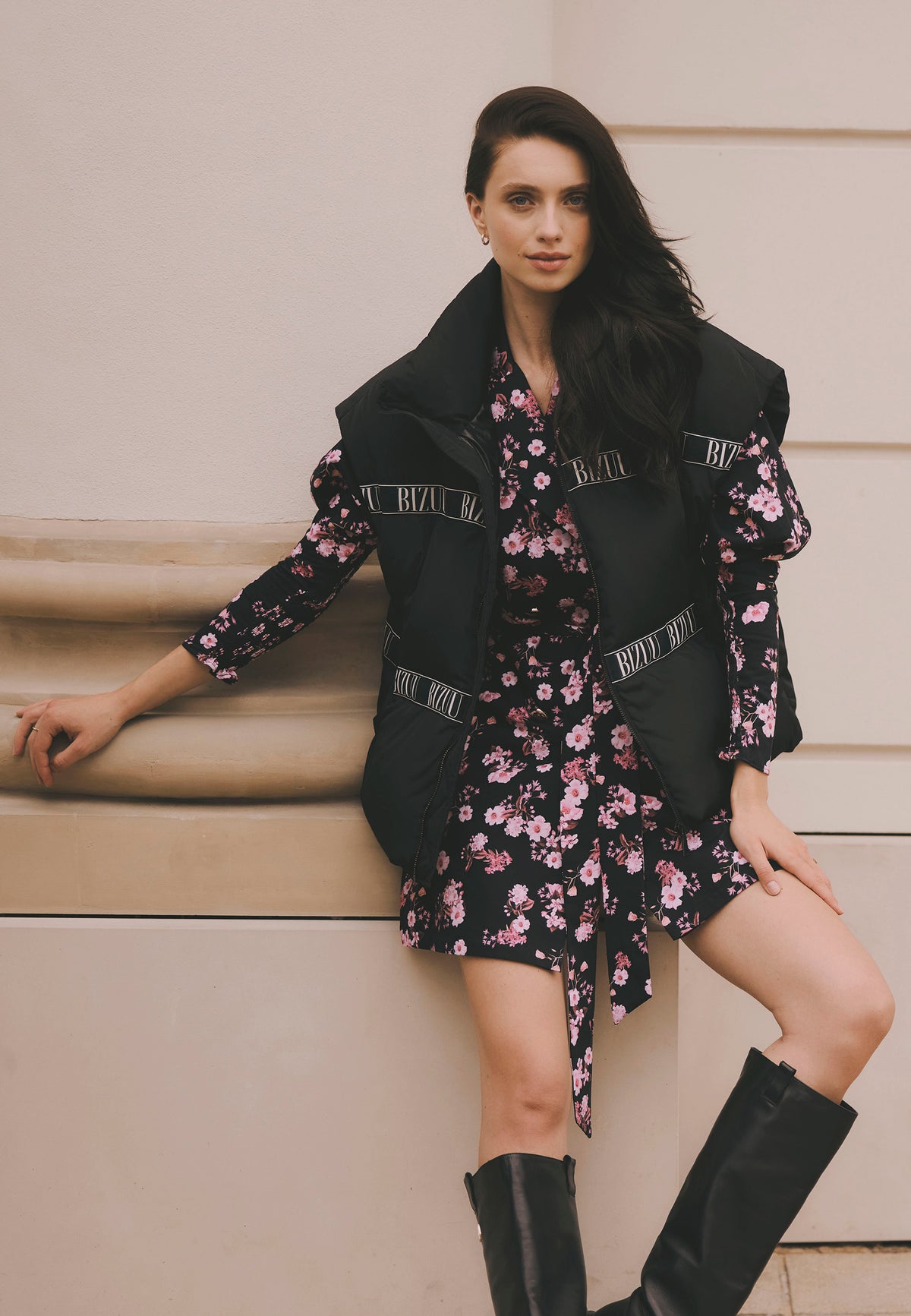 Model leaning against a building wall with one arm stretched out, dressed in the colorful GEORGINA dress accentuating the waist, paired with the AYASE down vest.