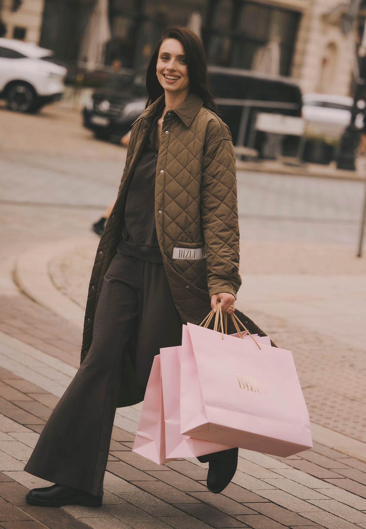 Model crossing the street with Bizuu shopping bags, dressed in the khaki MAREVIA maxi quilted coat.