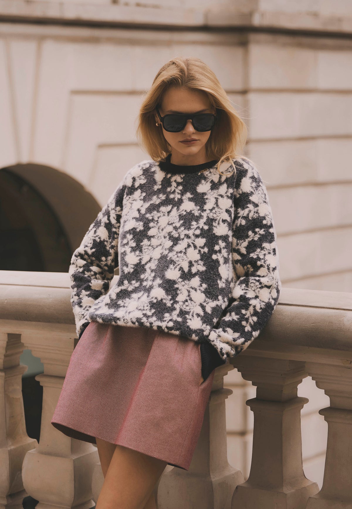 Model leaning against a wall with hands in the pockets of the burgundy BREDA mini skirt, styled with the floral CALTON sweater.