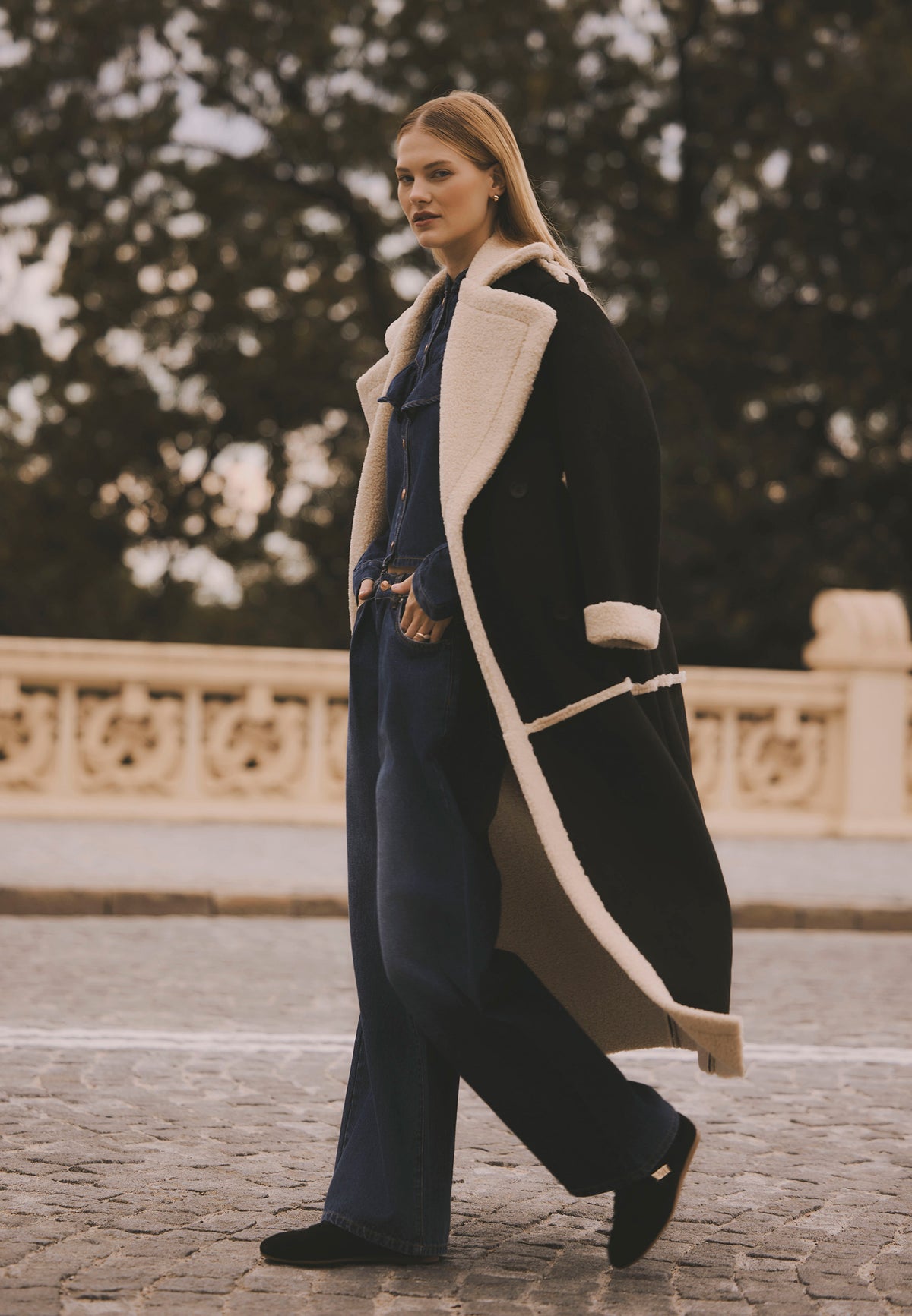 Model walking down the street, wearing the long black MAFARA shearling coat styled with the navy DILETTA shirt and LEYLA jeans.